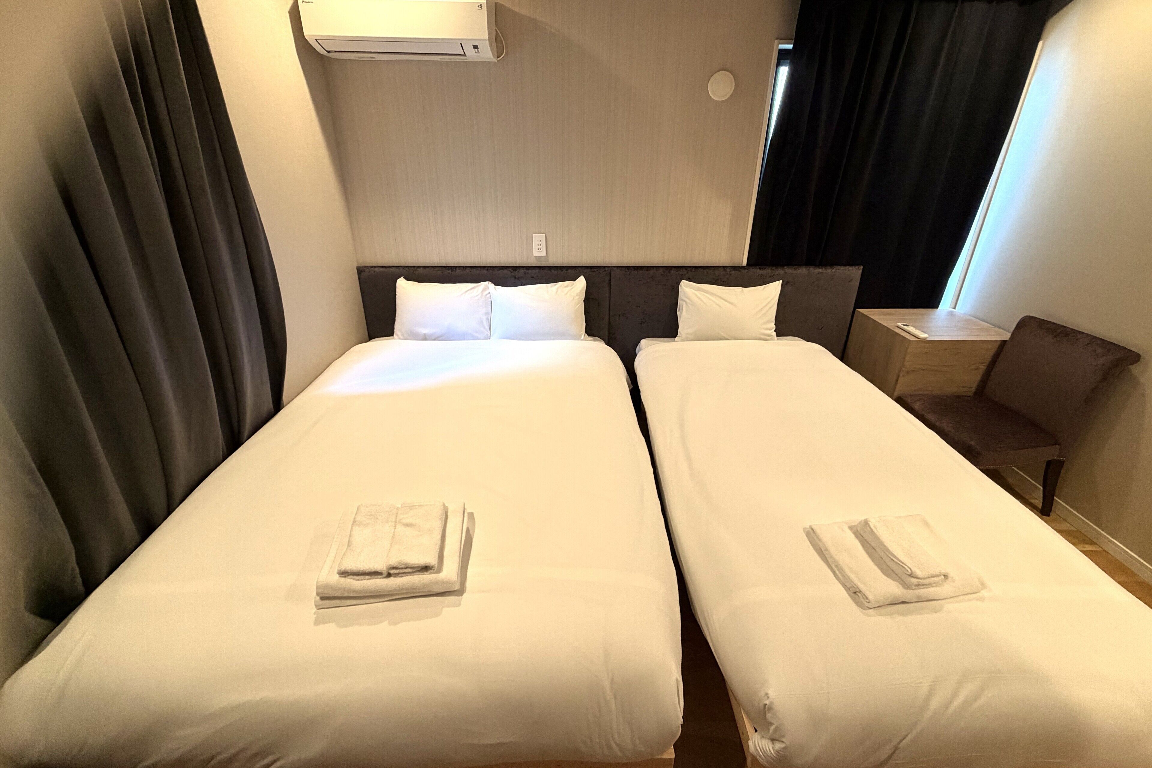 Een hotelkamer met twee bedden, elk voorzien van netjes opgevouwen handdoeken, een nachtkastje en een raam met gordijnen.