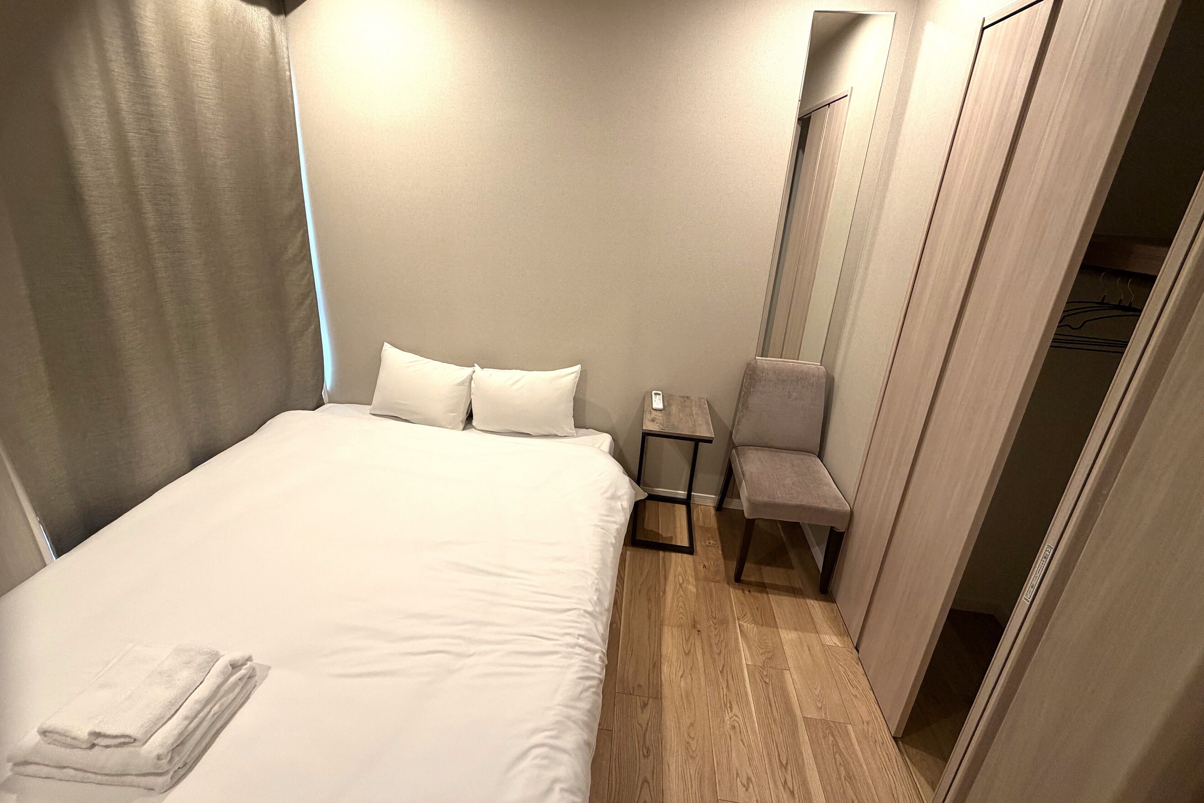 A neatly made bed with white linens, a small bedside table with a lamp, and a chair in a corner of a room with wooden flooring and a wardrobe.