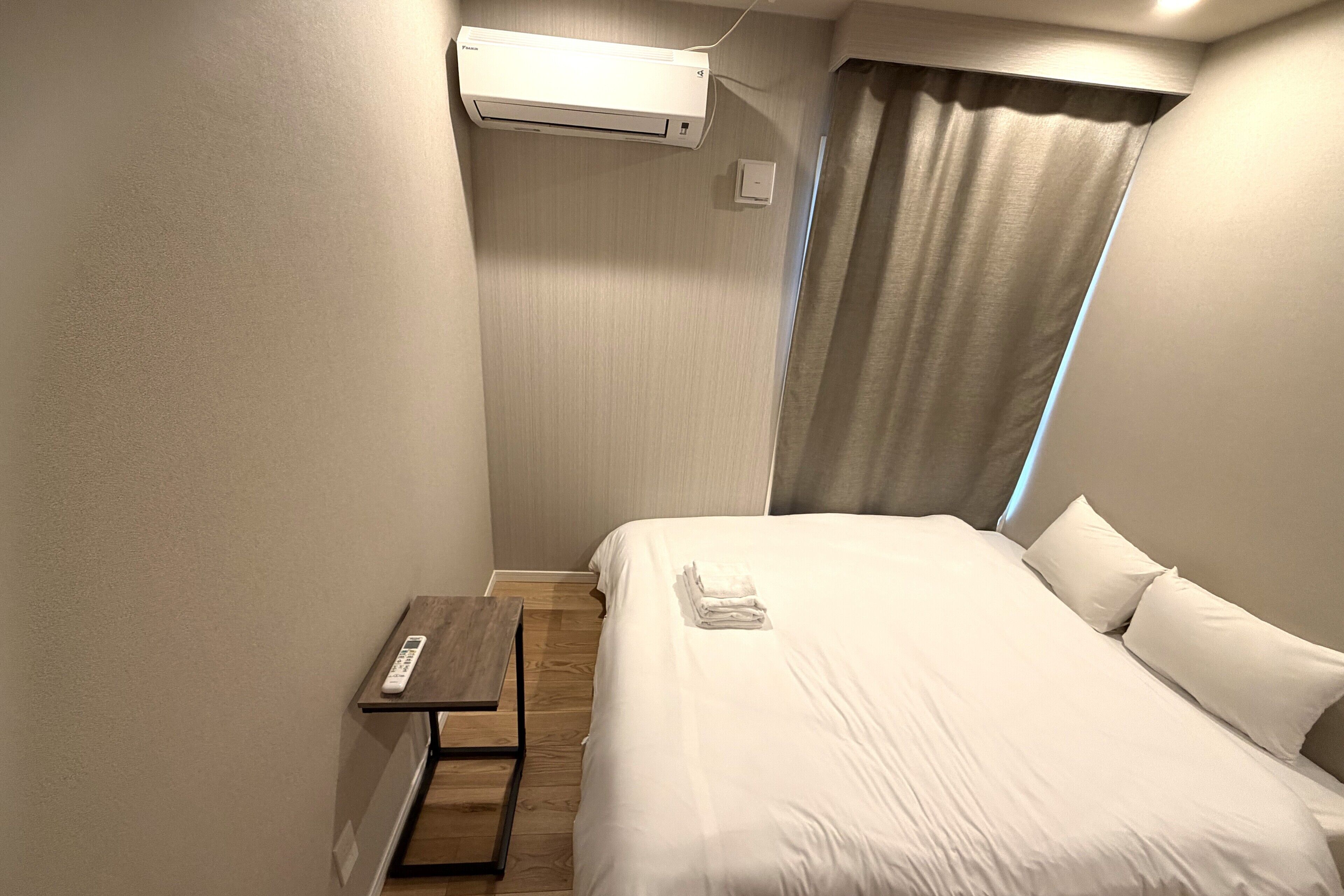 Habitación de hotel con una cama, una mesita de noche con un control remoto, un equipo de aire acondicionado y una cortina.