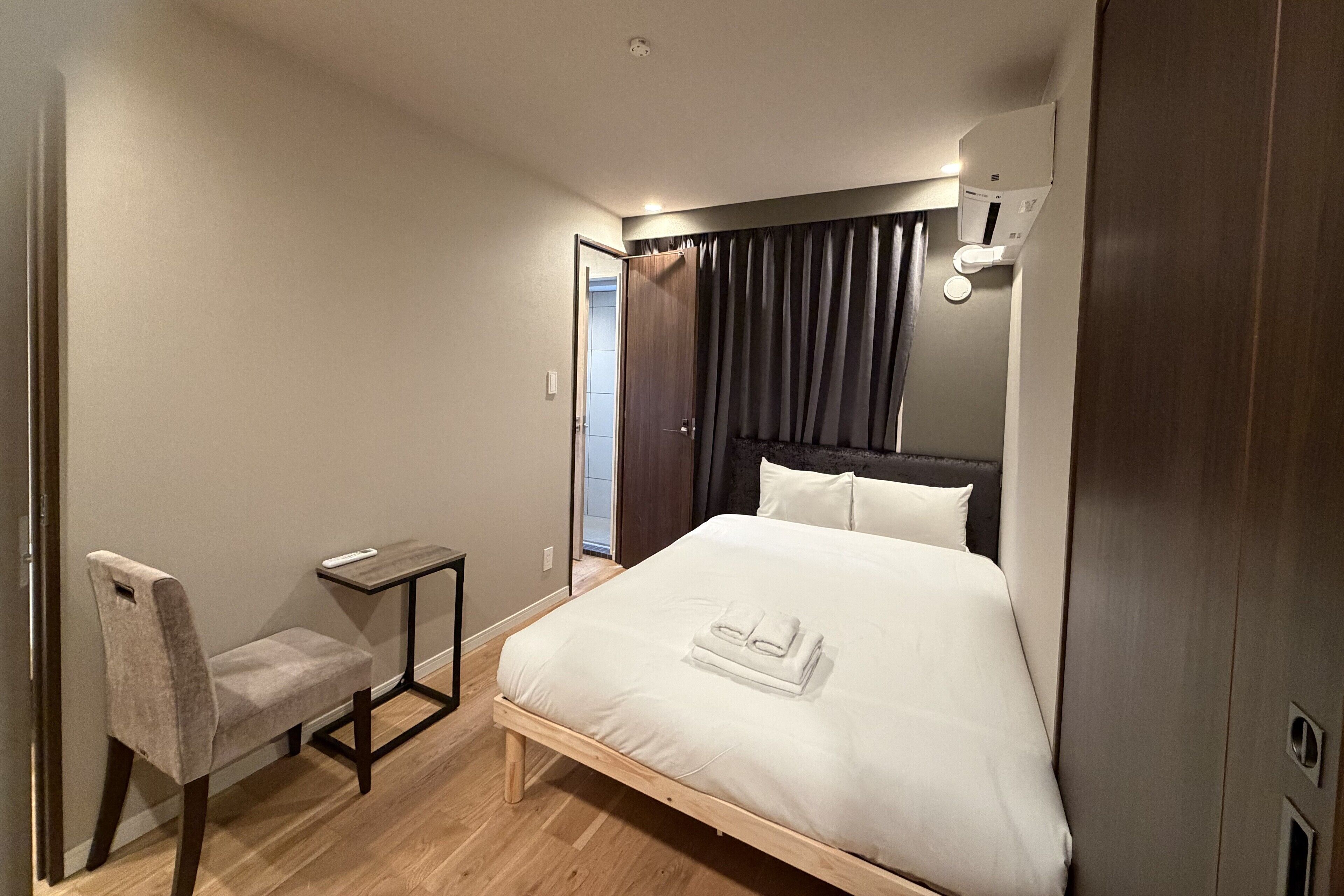 A neatly arranged bedroom with a bed, a chair, a small table, and a door leading to a bathroom.