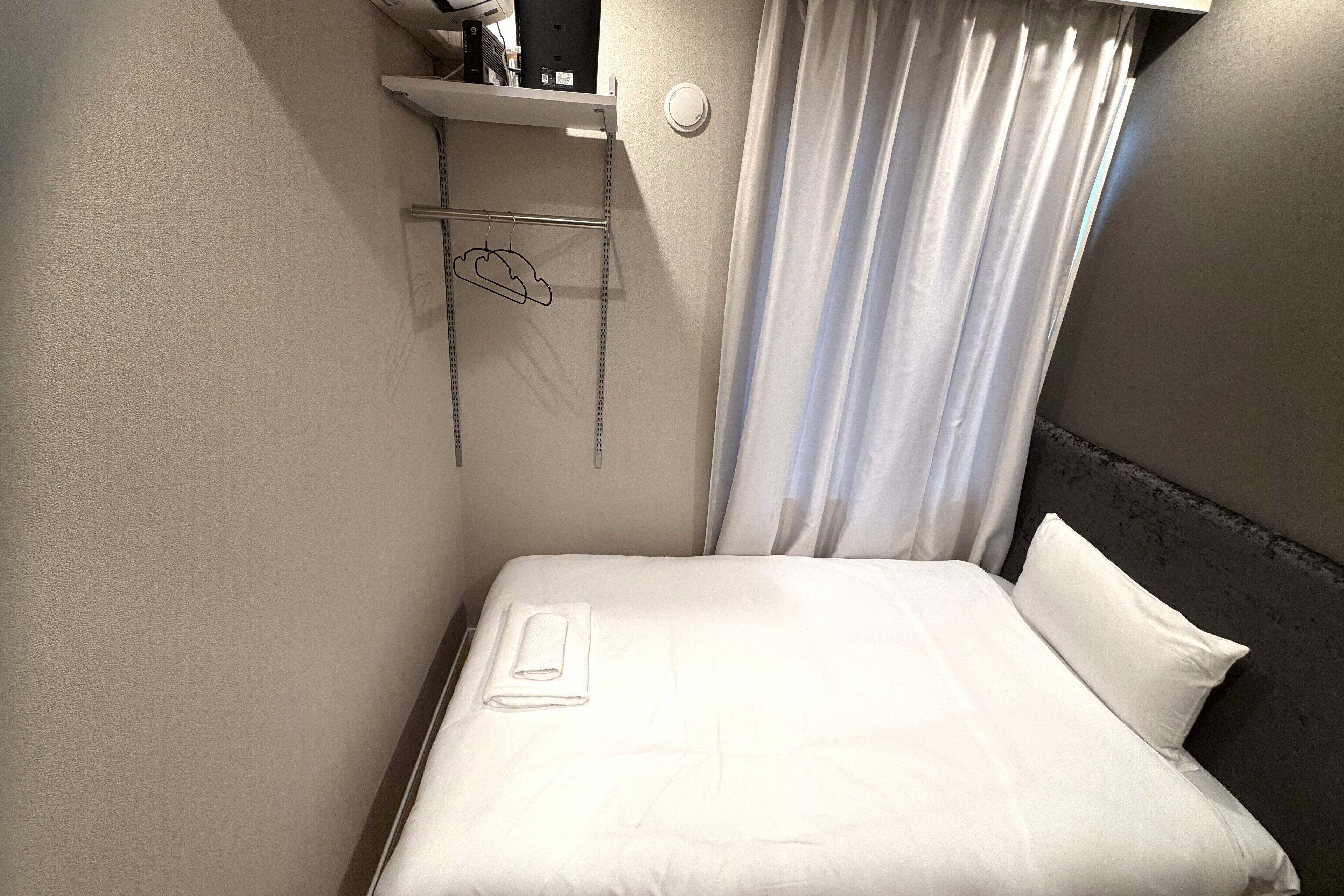 A neatly made bed with white linens, a folded towel, and a headboard. There is a shelf above the bed with a few items and a hanging coat rack with a cloud-shaped hanger.