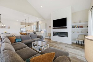 A modern living room with a sectional sofa, a flat-screen TV mounted on the wall, a fireplace, and a dining area in the background. - 5682 - Beers and Sunshine a Oceanside 5BR home in Nags Head, perfect for families and beach lovers! (Nags Head)