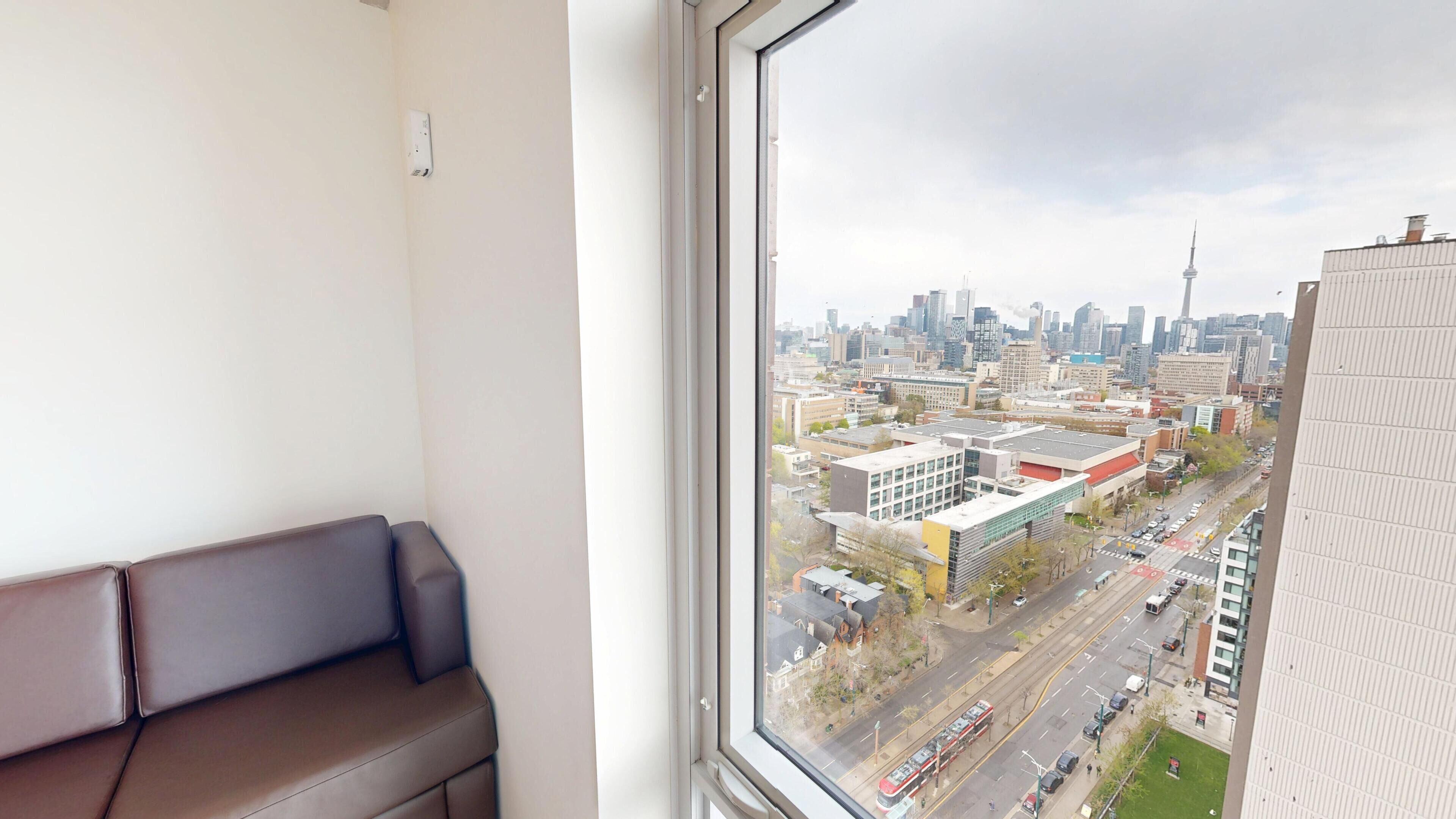 A room with a large window, a brown sofa, and a view of a cityscape with buildings and a tower.