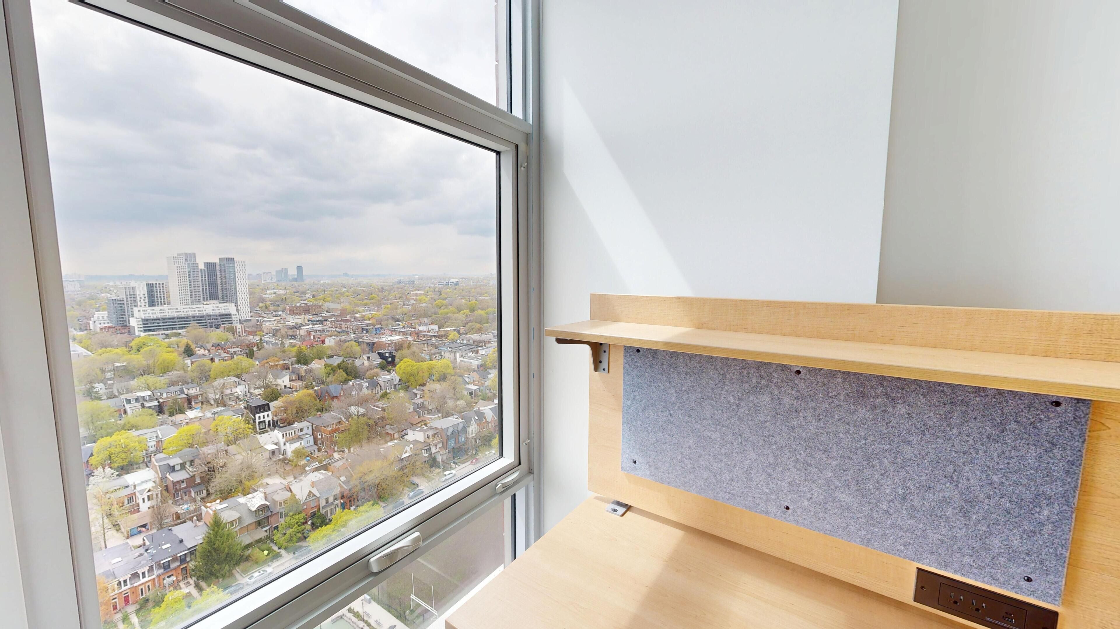 A room with a large window, a wooden desk, and a grey wall panel.