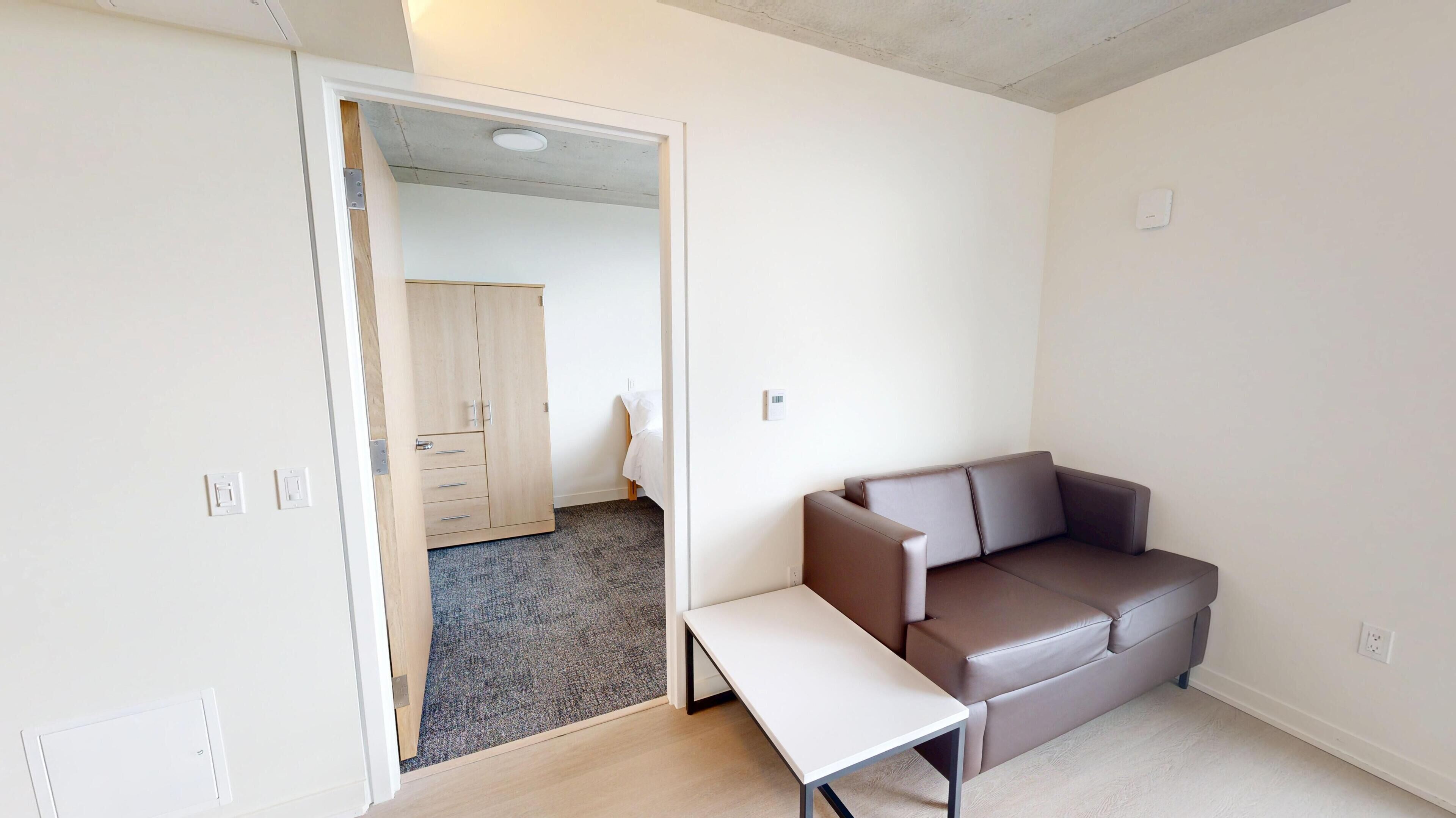 Een kamer met een bruine bank, een witte bijzettafel en een deur naar een andere kamer.