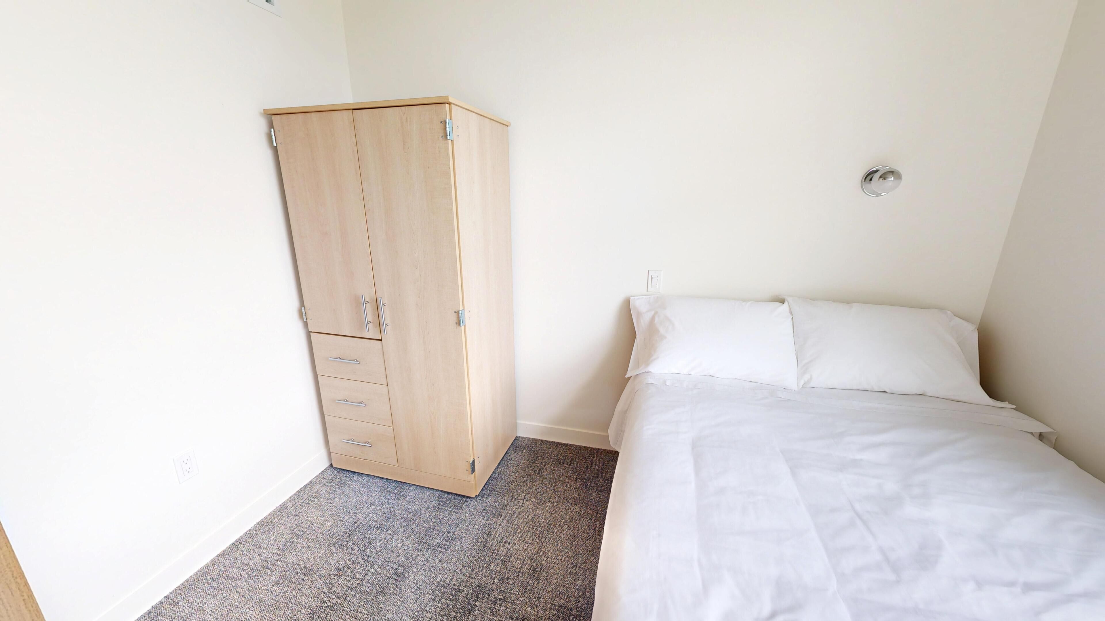A bedroom with a bed, a wooden wardrobe, and a carpeted floor.