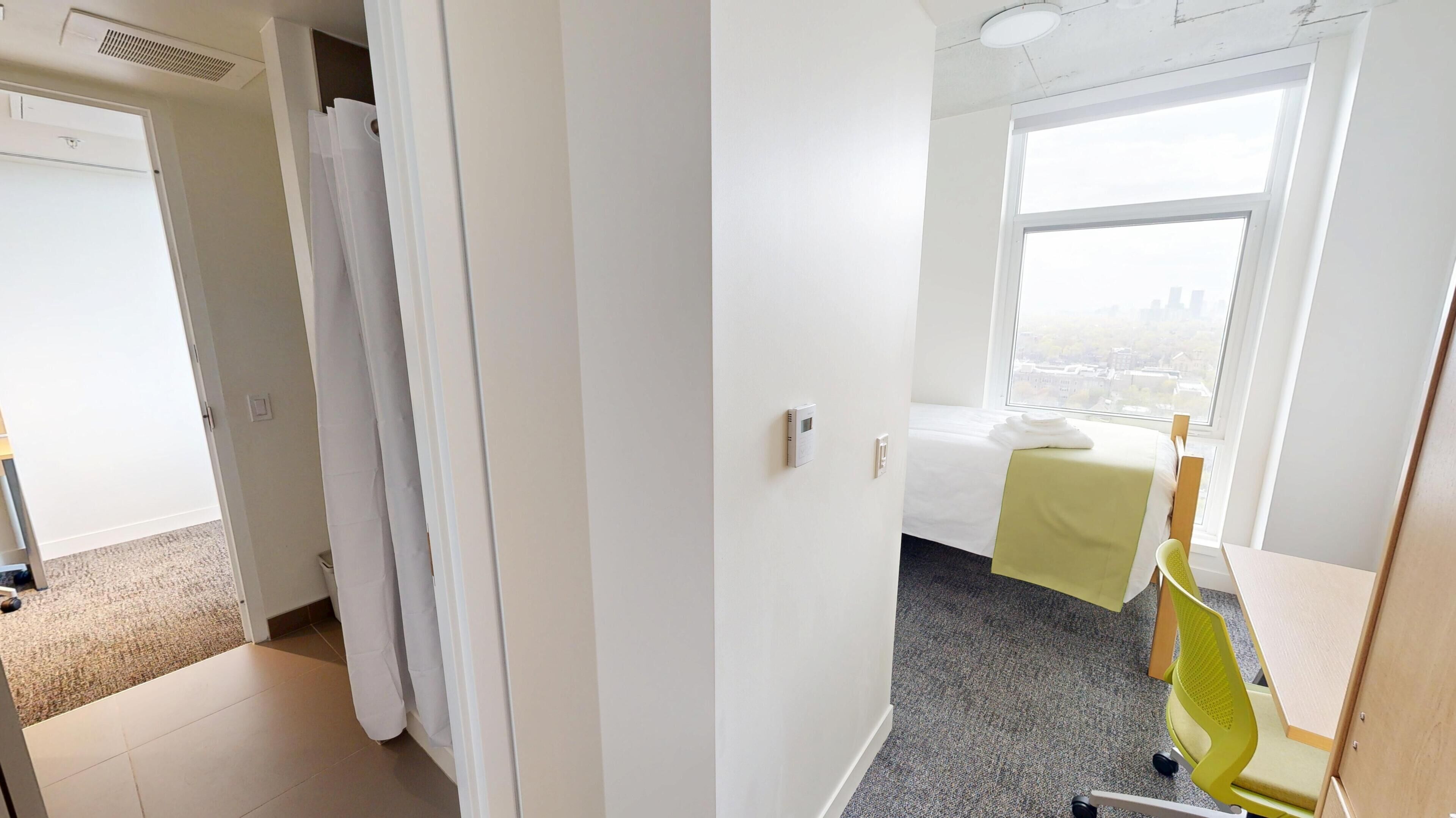 Une chambre d’hôtel comprenant un lit, une fenêtre donnant sur la ville, un bureau avec une chaise et une porte menant à une autre pièce.