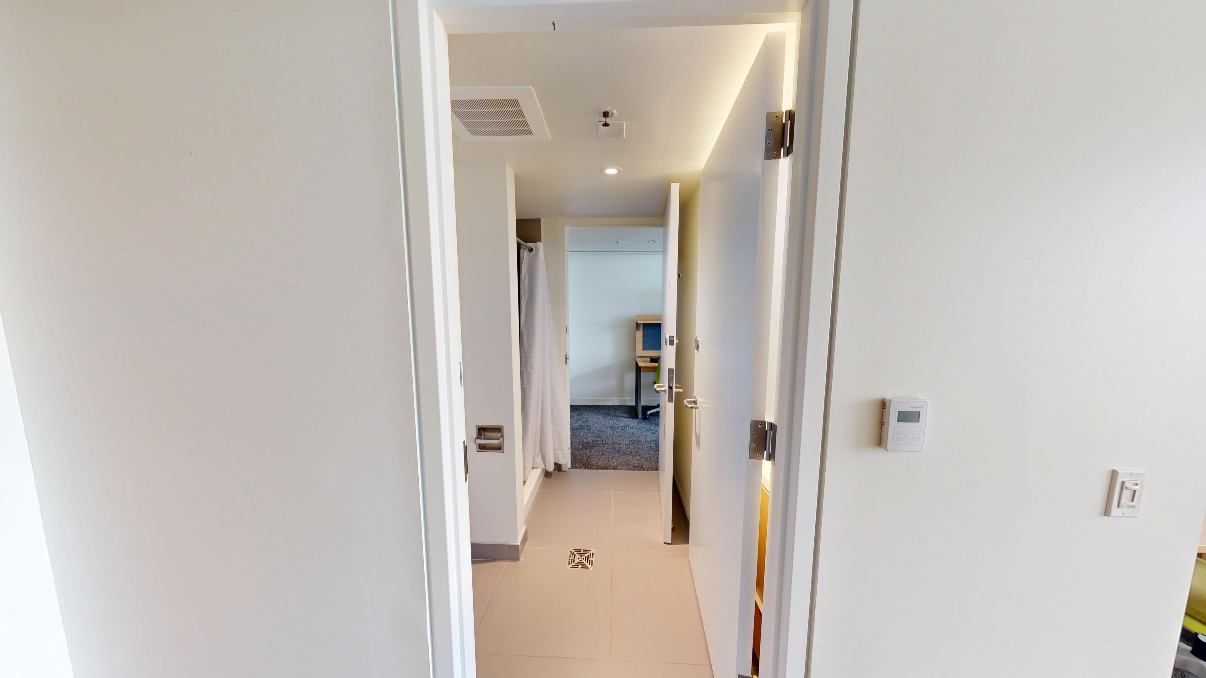 Een hotelgang met witte muren, een zichtbaar deurklink, een ventilatie rooster in het plafond en een lichtschakelaar aan de muur.