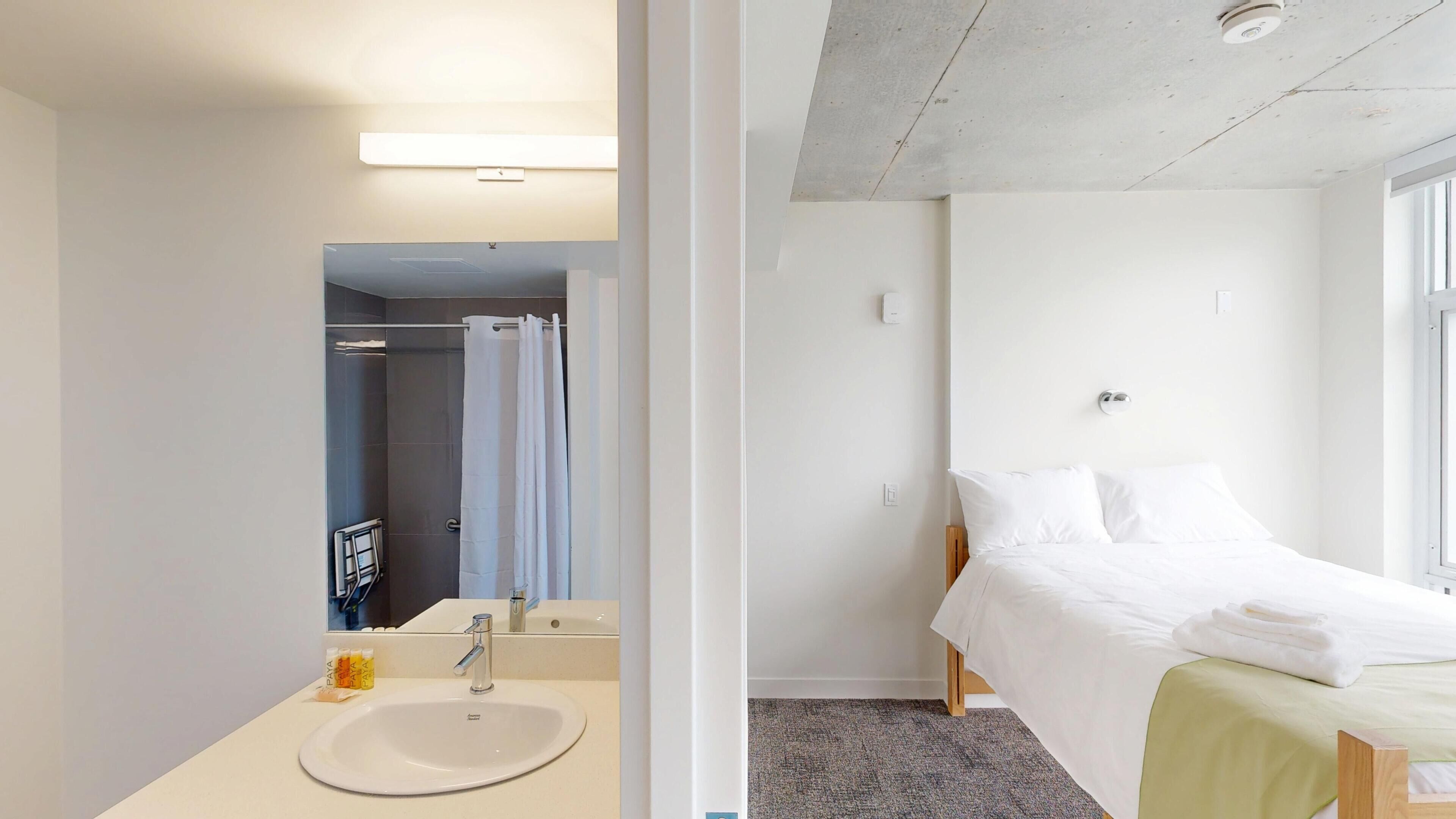 Une chambre d’hôtel avec un lit, une salle de bain comprenant un lavabo et un miroir, ainsi qu’une douche.
