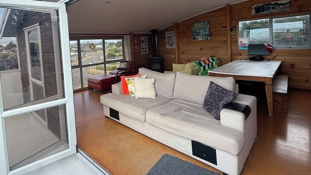 A modern living room with a large sofa, a dining table, and a television. - Waitarere Beach - panoramic beach views. Paradise in Summer & Winter (Levin)