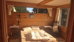 A wooden cabin bedroom with a bed, two bedside tables, a chair, and a window with curtains. - Waitarere Beach - panoramic beach views. Paradise in Summer & Winter (Levin)