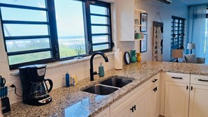 Cozinha com bancadas de granito, armários brancos, cafeteira e vista para o oceano através de grandes janelas.