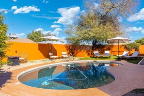 Une piscine entourée de chaises longues et de parasols, le tout bordé de murs orange et d’arbres.