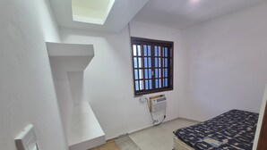 Ein kleiner, sauberer Raum mit einem Bett, einem Fenster mit Gitterstäben, einem Heizkörper und einem Lichtschalter an der Wand.
