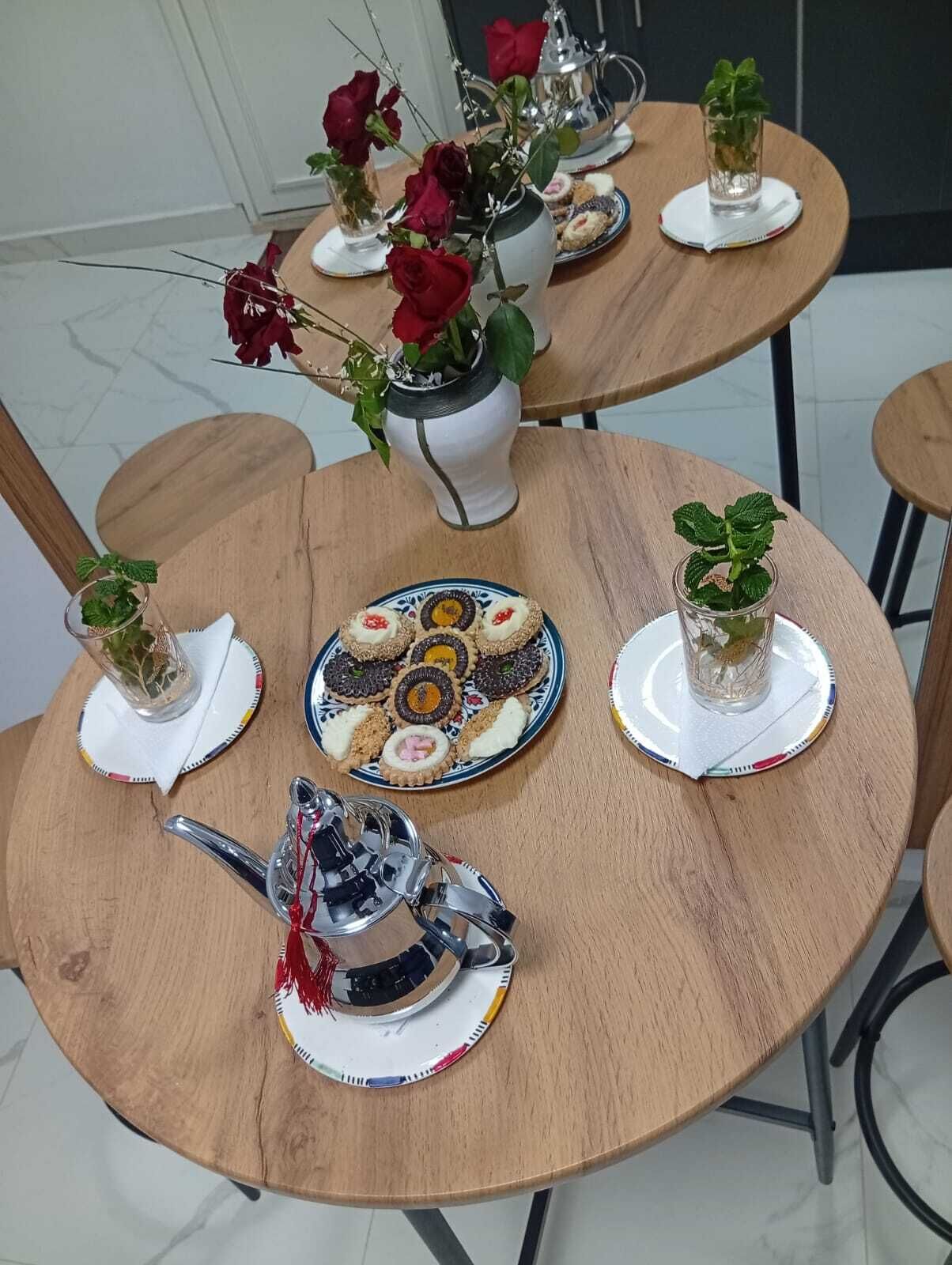 A round wooden table with a plate of assorted cookies, a vase with red roses, and small glass vases with mint plants.