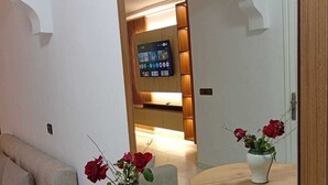 Ein modernes Wohnzimmer mit einer Couch, zwei runden Holztischen mit Rosenvasen, einem großen Spiegel, der den Raum reflektiert, und einem an einer Holzwand montierten Fernseher.