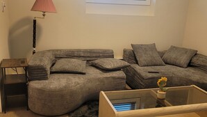 A living room with a sectional sofa, a glass coffee table, a lamp, and a window with blinds. - Beautiful new basement homeclose to greenery (Saskatoon)
