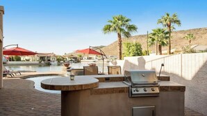 An outdoor barbecue area with a built-in grill, a seating area with a table and chairs, and a view of a body of water and palm trees.