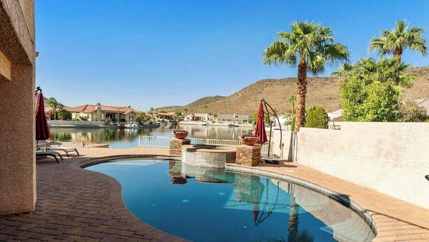 A swimming pool with a view of a lake and hills, surrounded by palm trees and lounge chairs.