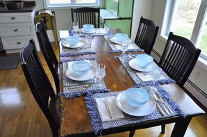 Una mesa de comedor con platos azules, vasos y cubiertos, rodeada de sillas negras en una habitación con piso de madera y una ventana.
