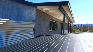 A modern building with a metal garage door, a covered porch with columns, and a view of mountains in the background. - Desert Game House with Hot Tub and Stargazing (Joshua Tree)