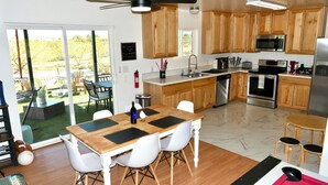 A modern kitchen with wooden cabinets, stainless steel appliances, and a dining area with a table and chairs. - Desert Game House with Hot Tub and Stargazing (Joshua Tree)
