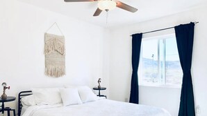A bedroom with a bed, two nightstands, a ceiling fan, a macrame wall hanging, and a window with curtains. - Desert Game House with Hot Tub and Stargazing (Joshua Tree)
