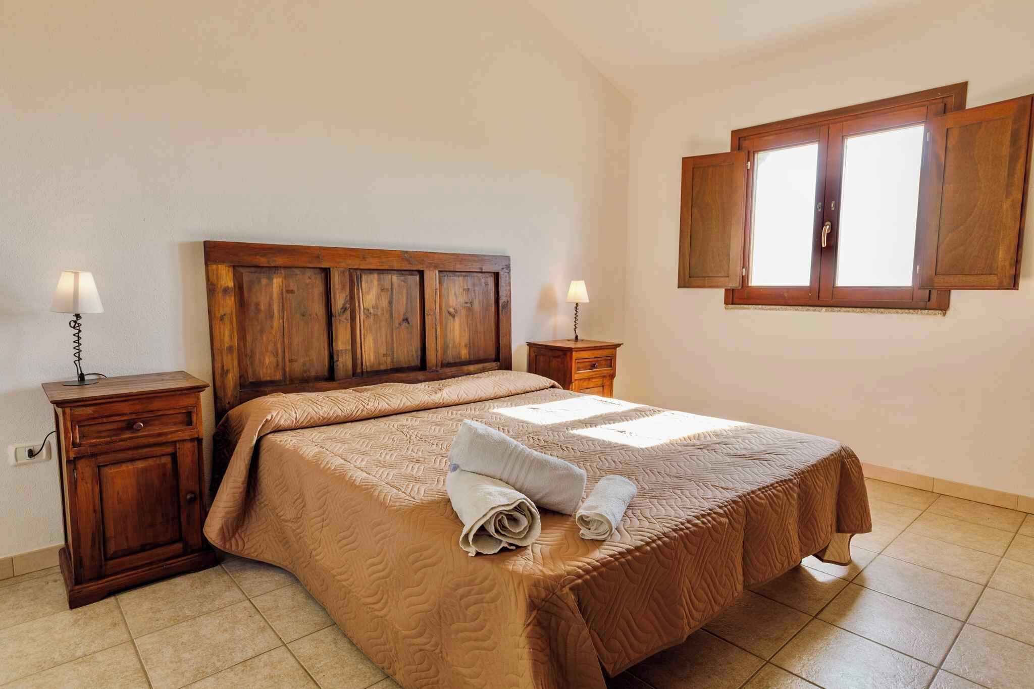 Un dormitorio con cabecera de madera, cama con colcha color beige, dos mesitas de noche con lámparas y una ventana con contraventanas de madera.
