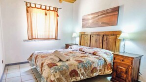 A bedroom with a wooden headboard, a large bed with a patterned quilt, a window with curtains, a bedside table with a lamp, and a painting on the wall. - Holiday Apartment Sea of Juniper Two-Room Apartment (Badesi)