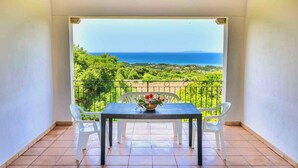 Varanda com mesa e cadeiras, com vista para uma paisagem deslumbrante com árvores e o mar.