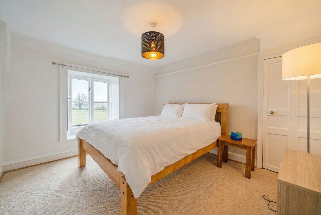 A bedroom with a wooden bed, a bedside table, a lamp, and a window with a view of the outdoors.