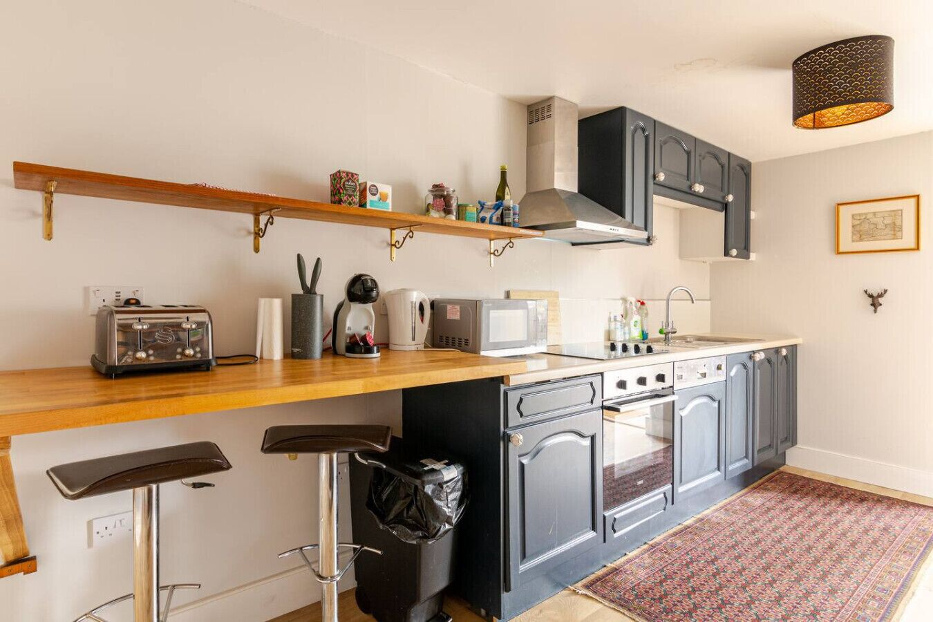 Une cuisine compacte avec un plan de travail en bois, un grille-pain, une bouilloire, un four à micro-ondes et un évier. On y trouve également des tabourets de bar, une poubelle et un tapis à motifs sur le sol.