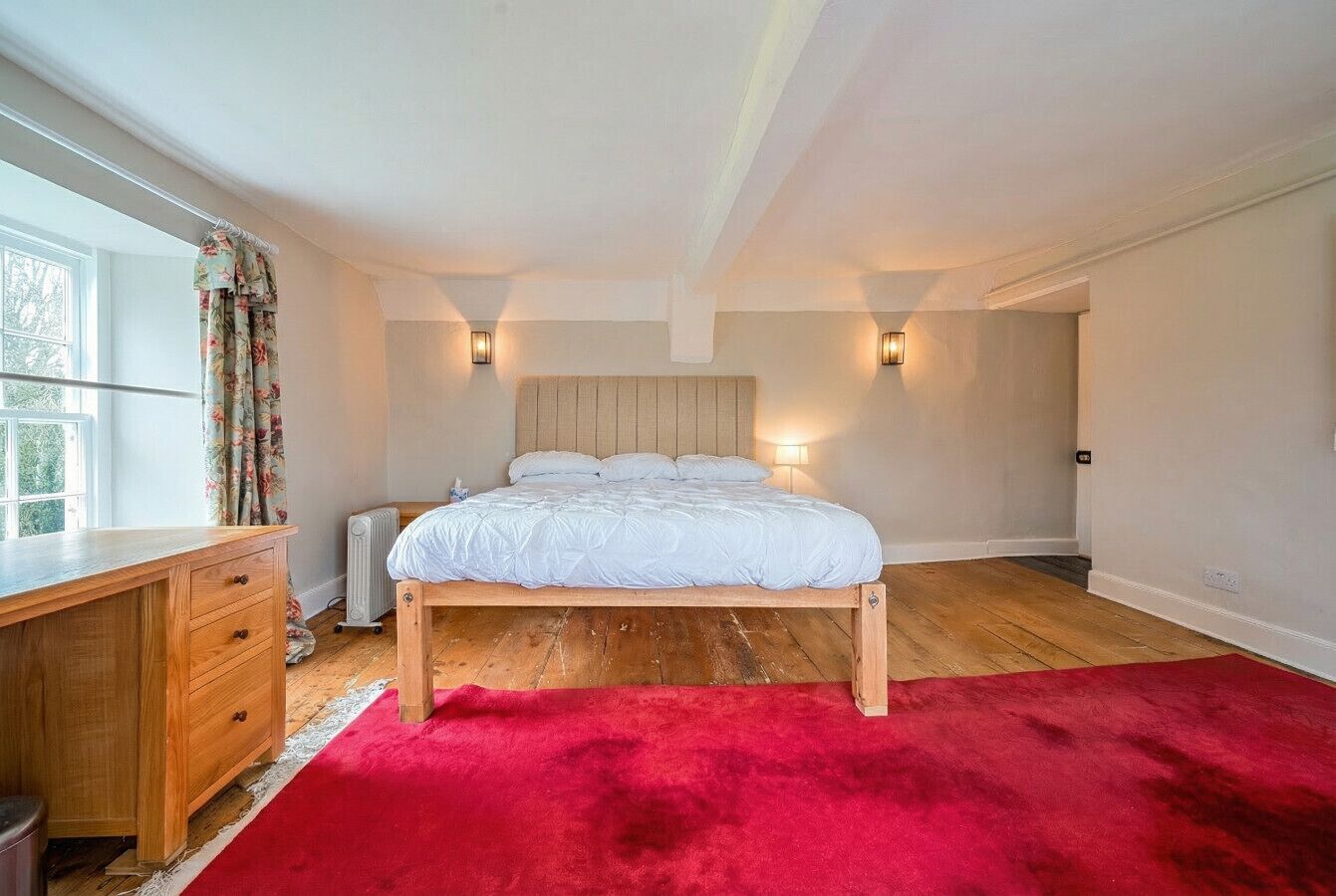 Une chambre avec un lit en bois, un tapis rouge, un bureau en bois et deux lampes fixées au mur.