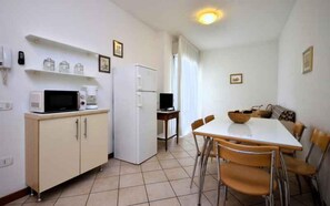 Eine kleine Küche mit Mikrowelle, Kaffeemaschine und Kühlschrank. Ein Esstisch mit Stühlen und einer Schüssel steht daneben.