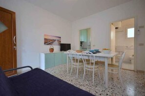 Quarto compacto com cama, mesa de jantar com cadeiras, pequena cozinha e um quadro na parede.