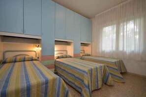 Quarto com duas camas, cada uma com roupa de cama listrada, armários claros azuis acima e uma grande janela com cortinas leves.