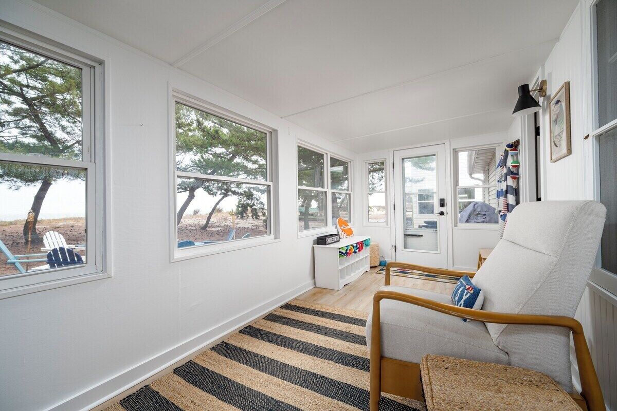 A bright, sunlit room with large windows, a striped rug, a white sofa, and a wooden chair.