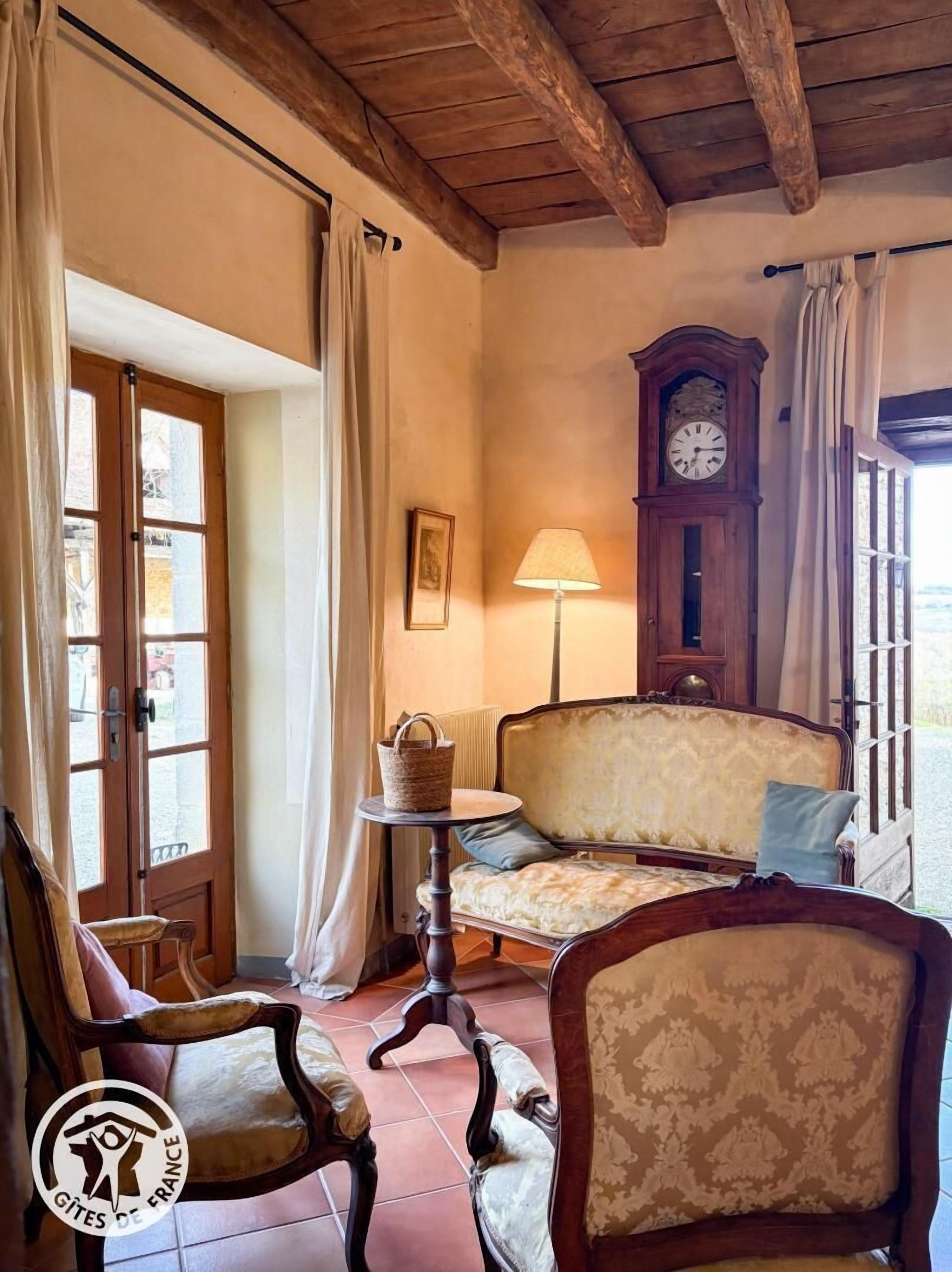A cozy room with a vintage sofa, a wooden chair, a small table with a lamp, a clock, and a door with curtains.