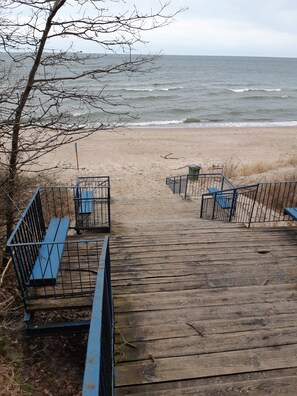 Ein hölzerner Steg führt zu einem Sandstrand mit Bänken und Geländern.