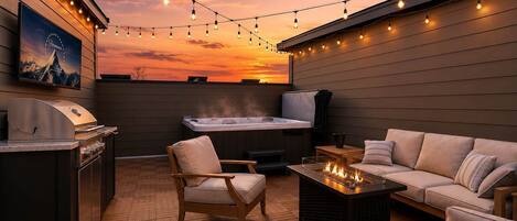 A rooftop patio with a grill, seating, a hot tub, and string lights against a sunset.