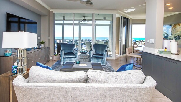 Een ruime woonkamer met een grijze bank, twee blauwe fauteuils en een salontafel. Er is een plafondventilator te zien, en de kamer heeft grote ramen met uitzicht op de oceaan.