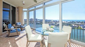 Een balkon met een glazen tafel, witte stoelen, rieten stoelen met blauwe kussens en uitzicht op de stad en het water.