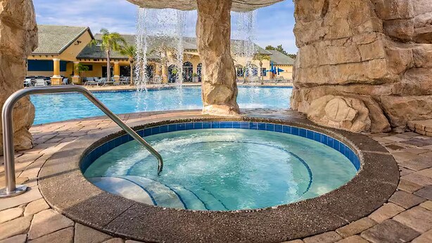 Un bain à remous surmonté d’une arche en pierre, une piscine avec des fontaines et, au fond, un bâtiment doté d’un toit en tuiles.