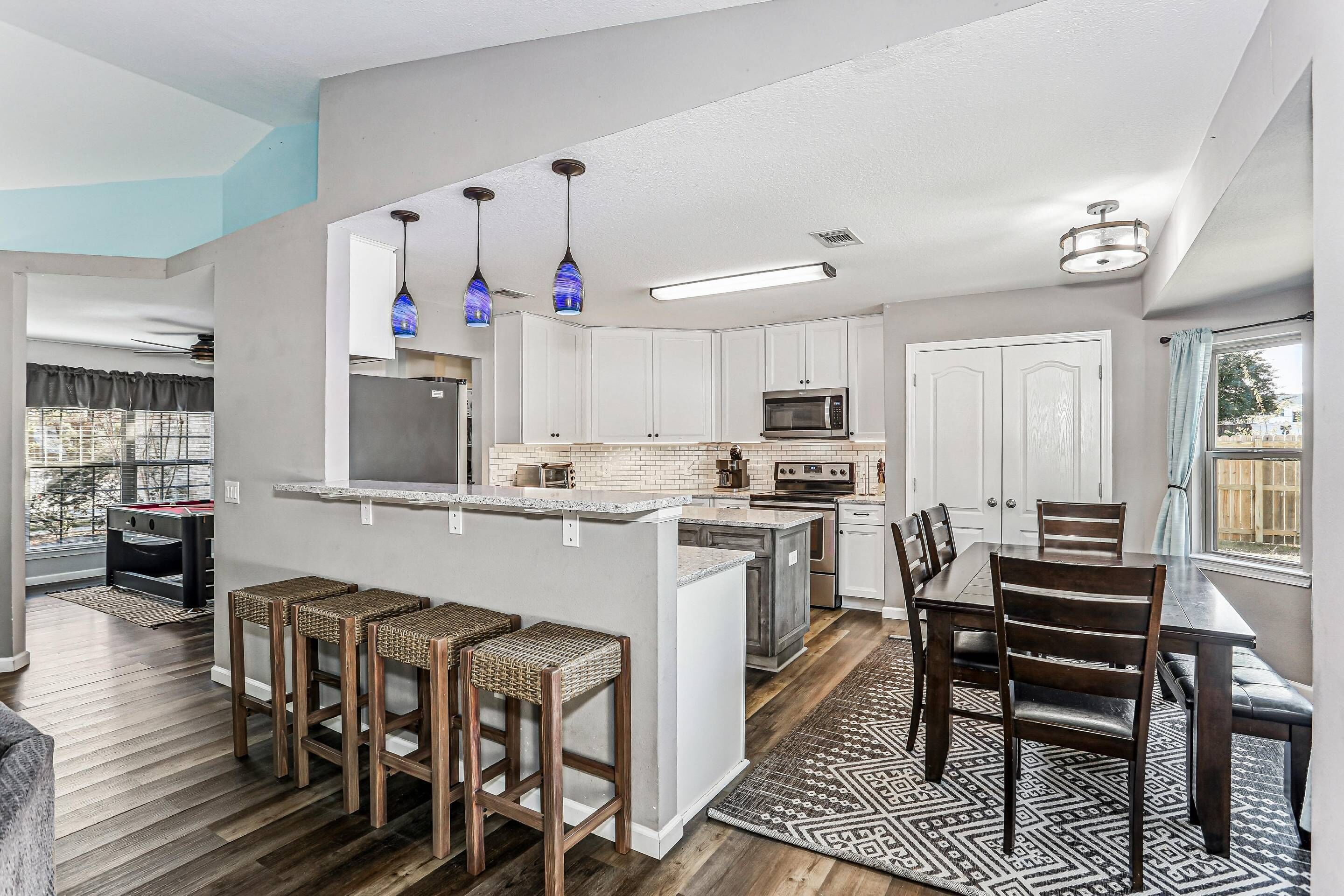 Une cuisine moderne avec des meubles blancs, un îlot central doté de tabourets, un coin repas avec une table et des chaises, et un tapis à motifs.