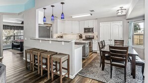 A modern kitchen with white cabinets, a central island with stools, a dining area with a table and chairs, and a patterned rug. - 4BR Home with Volleyball Court on a Huge Lot Just Minutes from Navarre Beach - Dog Friendly (Navarre)