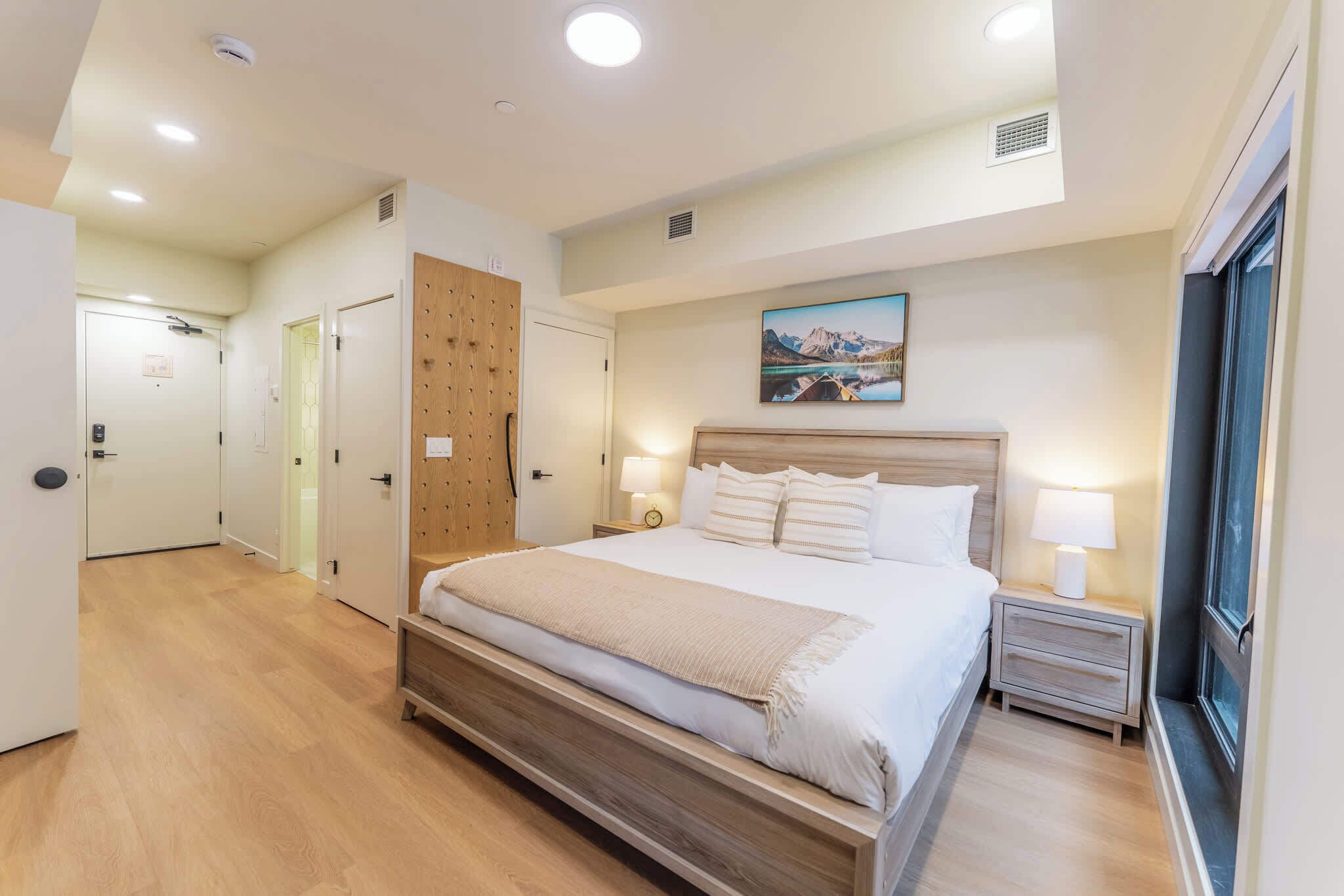 A modern hotel room with a large bed, bedside lamps, a wooden wardrobe, and a painting on the wall.