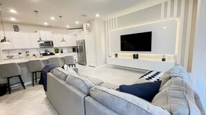 A modern kitchen with white cabinets, a built-in microwave, and a refrigerator. There is a dining area with a gray sofa and a flat-screen TV mounted on the wall. - 7545 OS - Solterra Resort Grandeur Luxurious 5-Bedroom Pool Home (Davenport)