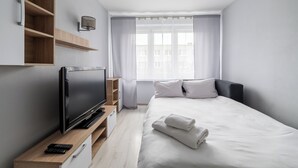 Una moderna camera d'hotel con una TV a schermo piatto, scaffali in legno, un comodino, un letto con lenzuola bianche e una finestra con tende trasparenti.