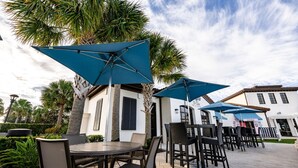 Patio esterno con tavoli e sedie rotonde, ombrelloni blu e palme.
