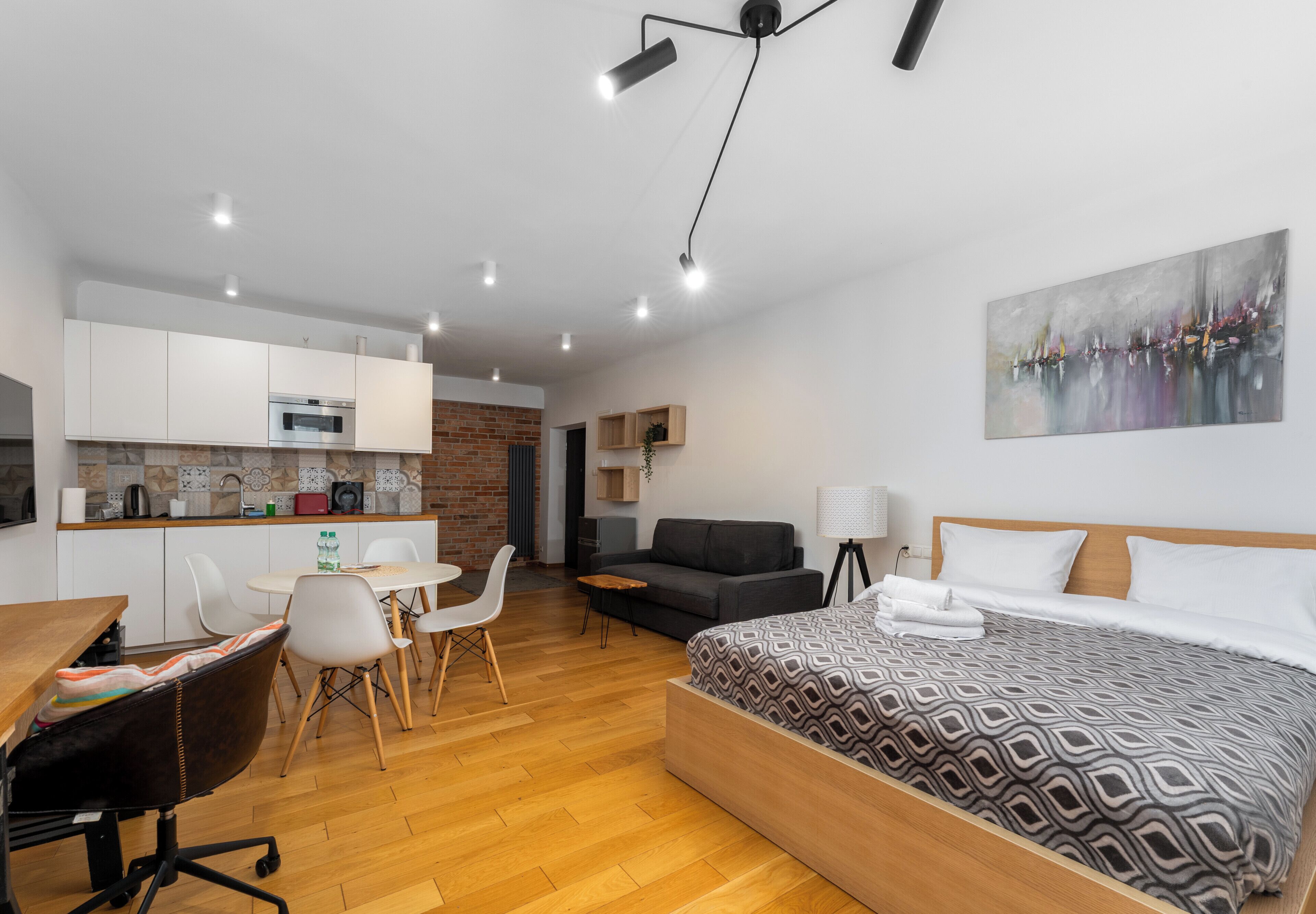 A modern hotel room with a wooden bed, a desk, a dining area, and a kitchenette.